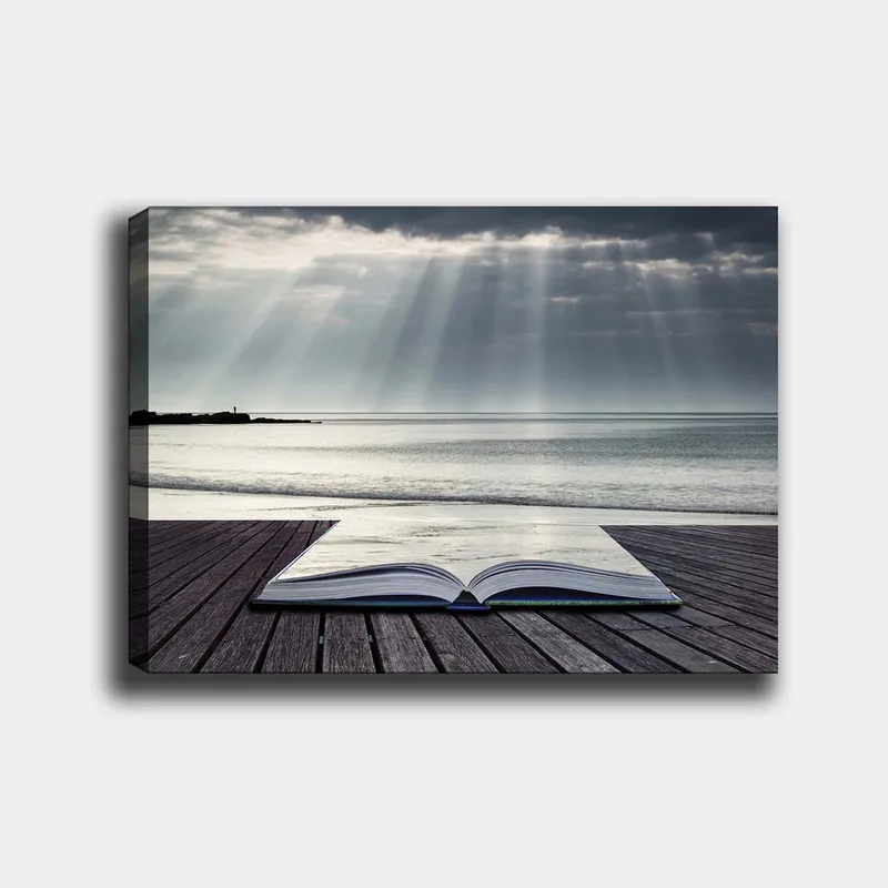 Canvastavla 70x100 cm - Öppen bok som ligger på en träbrygga vid havet, omgiven av mjuka ljusstrålar från en molnig himmel, Grå / Blå / Vit