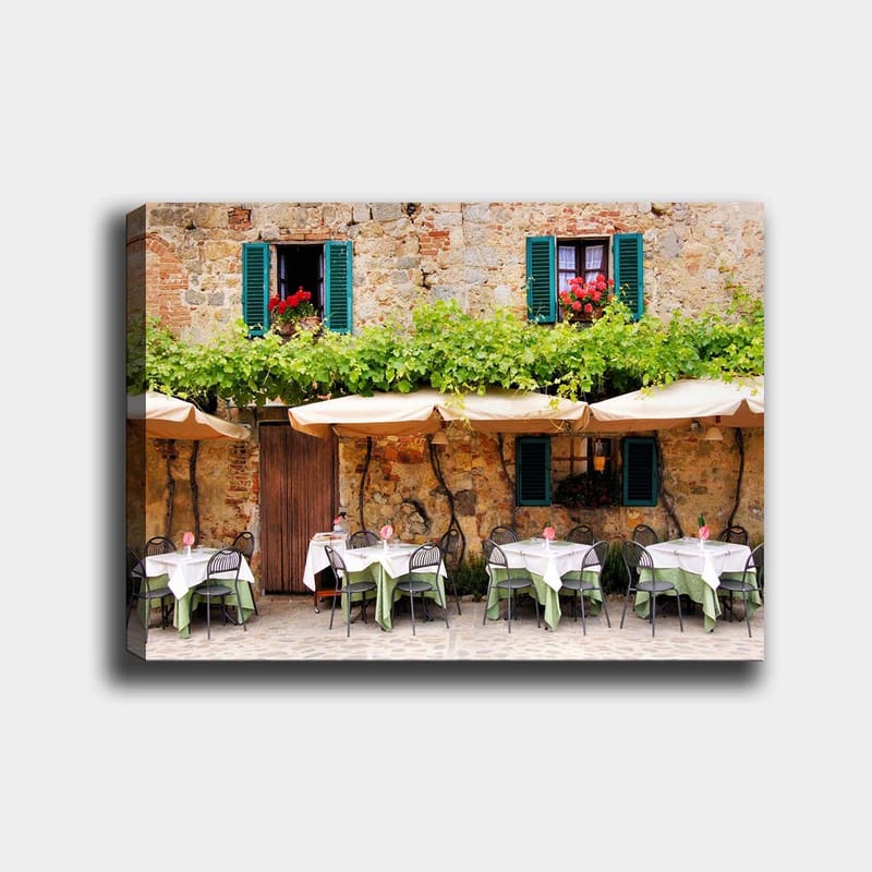Canvastavla 70x100 cm - Charmig uteservering med vita bord och gröna dukar framför en gammal stenbyggnad med fönsterluckor i mörkgrön färg - Mörkgrön / Vit / Tegelröd - Inredning - Tavlor & konst - Canvastavlor