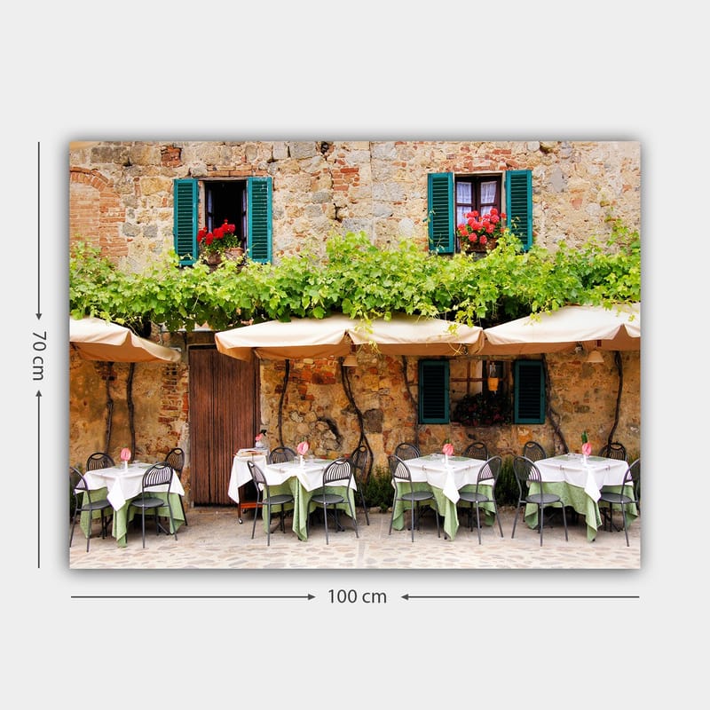Canvastavla 70x100 cm - Charmig uteservering med vita bord och gröna dukar framför en gammal stenbyggnad med fönsterluckor i mörkgrön färg - Mörkgrön / Vit / Tegelröd - Inredning - Tavlor & konst - Canvastavlor