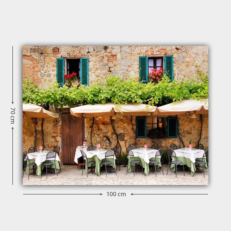 Canvastavla 70x100 cm - Charmig uteservering med vita bord och gröna dukar framför en gammal stenbyggnad med fönsterluckor i mörkgrön färg - Mörkgrön / Vit / Tegelröd - Inredning - Tavlor & konst - Canvastavlor
