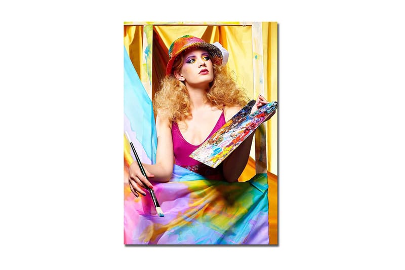 Canvastavla 70x100 cm - Kreativ kvinna som målar med en palett i handen, omgiven av färgglada tyger, Rosa / Blå / Gul