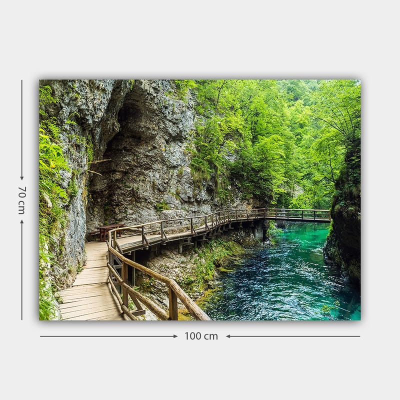 Canvastavla 70x100 cm - Naturskön vy av en träbro som slingrar sig genom en grönskande ravin vid en klar, turkos flod - Grön / Turkos / Grå - Inredning - Tavlor & konst - Canvastavlor